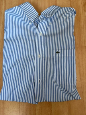 Blårandig skjorta från Lacoste - Snygg blårandig skjorta från Lacoste med korta ärmar och klassisk krage. Skjortan har knäppning framtill och en bröstficka med Lacostes ikoniska krokodillogga. Storlek 38 (S).
