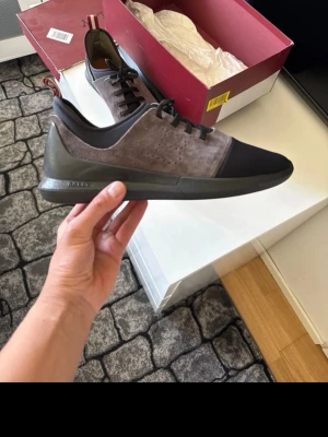 Bally  sneakers i brun(gråt) och svart - Stilrena sneakers från Bally mocka med svarta detaljer och tåparti. Skorna har svarta snören, diskret perforering på sidan och en mörk sula. Perfekta för dig som gillar en clean och modern look.