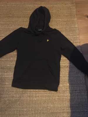 Svart hoodie från Lyle & Scott - Snygg svart hoodie från Lyle & Scott med klassisk gul logga broderad på bröstet. Perfekt för en avslappnad och stilren look.