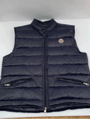 Mörkblå väst från Moncler - Snygg mörkblå dunväst från Moncler med klassisk quiltad design och dragkedja framtill. Västen har två fickor med dragkedja och Monclers ikoniska logotyp på bröstet. Perfekt för lager-på-lager och streetstyle.