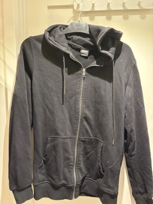 Svart zip hoodie från Urban Classics | S - Svart hoodie med dragkedja från Urban Classics. Klassisk modell med huva, dragsnören och två stora fickor framtill. Tillverkad i mjukt bomullsmaterial som är skönt att ha på sig. Perfekt för en avslappnad streetstyle-look.