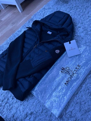Svart pufferjacka med huva från Moncler - Snygg svart pufferjacka från Moncler med huva och stickade ärmar. Jackan har dragkedja framtill, en ficka med dragkedja på ärmen och klassisk Moncler-logga. Perfekt för kalla dagar och riktigt trendig look.