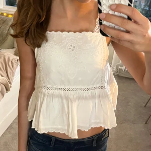 Söt topp med volang - Supersöt vit linnetopp med broderade detaljer och volang nertill. Toppen har smala axelband, fyrkantig ringning och spetskant upptill och nertill.