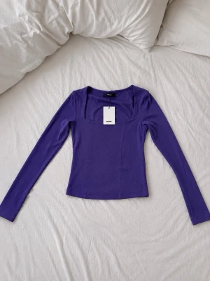 Lila långärmad topp från NA-KD - Trendig lila topp från NA-KD med lång ärm och fyrkantig ringning. Toppen är figurnära och har en ribbad struktur som ger en snygg och modern look. Perfekt att styla med både jeans och kjol för en cool vibe.