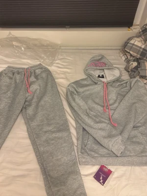 Syna world  - Mysigt loungewear-set i ljusgrått med matchande hoodie och byxor. Hoodien har rosa dragsko och tryck i luvan, samt känguruficka. Byxorna har resår i midjan och rosa dragsko. Perfekt för chill dagar hemma.