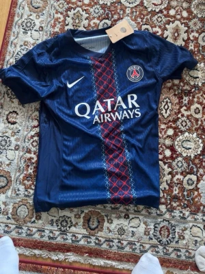 PSG matchtröja Nike blå - Snygg blå PSG matchtröja från Nike med Qatar Airways-tryck på bröstet och Paris Saint-Germain-logga. Tröjan har röd och vit detalj i mitten, korta ärmar och spelarnamn D. Doué samt nummer 14 på ryggen. Tillverkad i lätt och ventilerande material. 