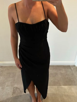 Zara Svart långklänning med öppen rygg - Snygg svart långklänning med tunna axelband och en smickrande omlott-detalj framtill. Klänningen har öppen rygg med knyt och en elegant slits framtill. Perfekt för dig som vill ha en stilren och trendig look. Köpte ny på Zara för 2 år sedan men är helt oanvänd! 