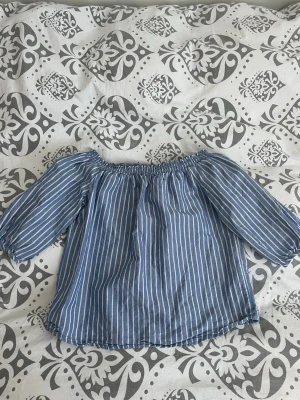 Randig offshoulder blus i blått - Snygg blåvit randig blus med offshoulder-modell och trekvartsärm. Blusen har resår vid axlarna och är tillverkad i ett lätt och luftigt tyg.