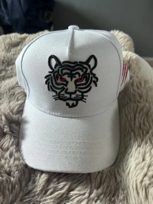 Vit keps med tigerbrodyr - Snygg vit keps med broderad tiger i svart och rött framtill samt röda klösmärken på sidan. Justerbar baktill och tillverkad i bomullstyg. Perfekt för dig som vill sticka ut med en cool detalj.