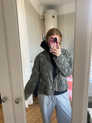 Grön camouflage vindjacka - Snygg grön vindjacka i camouflage-mönster med knappar framtill och flera fickor. Jackan har en avslappnad passform och är perfekt för dig som gillar streetwear-vibe. Tillverkad i bomull och polyester för en bekväm känsla.