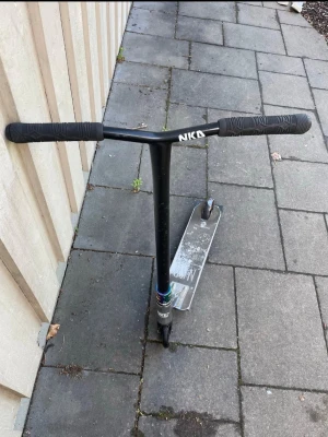 Svart trick sparkcykel från NKD rally v4 - Säljer en svart trick sparkcykel från NKD med breda handtag och robust metallram. Styret är svart med greppvänliga handtag och decket är i metall med NKD-logga. Perfekt för skateparken eller att cruisa runt på stan.