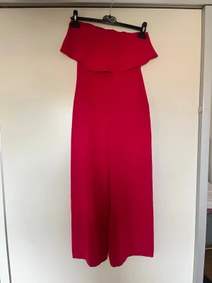 Röd/Rosa off shoulder jumpsuit - Snygg röd rosa jumpsuit med off shoulder-design och vida ben. Jumpsuiten har en volang upptill som ger en trendig look. Perfekt för dig som vill sticka ut med färg och stil. Materialet känns mjukt och följsamt, troligtvis polyester eller liknande. Silikon vid bröstet så att det inte glider ner oanvänd 