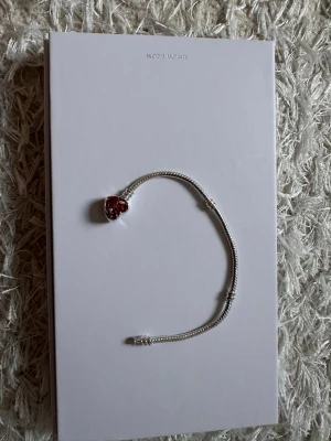 Pandora armband med hjärtberlock - Snyggt silverfärgat armband från Pandora med en hjärtformad berlock. Berlocken har röda och silvriga detaljer som ger en romantisk vibe. Perfekt att matcha med andra smycken eller bära som statement piece.