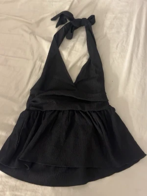 Svart halterneck topp med knyt - Säljer en svart halterneck topp med knyt i nacken och v-ringad front. Toppen har en lätt rynkad, flowy nederdel och är tillverkad i ett strukturerat tyg som ger en snygg look. Perfekt för dig som vill ha en stilren och trendig topp.