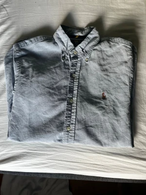 Ljusblå skjorta från Polo Ralph Lauren - Klassisk ljusblå skjorta från Polo Ralph Lauren med broderad logga på bröstet. Skjortan har knappar framtill och button-down krage. Perfekt för en stilren look och tillverkad i mjuk bomull.