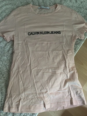 Ljusrosa Calvin Klein Jeans t-shirt - Säljer en ljusrosa t-shirt från Calvin Klein Jeans med svart logga framtill. Klassisk rund halsringning och korta ärmar. Tillverkad i mjuk bomull för en skön känsla. Perfekt till jeans eller shorts för en clean och enkel look.