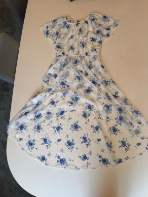 Vit långklänning med blå blommor - Superfin långklänning i vitt med blått blommönster. Klänningen har korta ärmar och en luftig, flowy passform som ger en romantisk vibe. Perfekt för varma dagar och somriga tillfällen. Tillverkad i ett lätt och mjukt material.