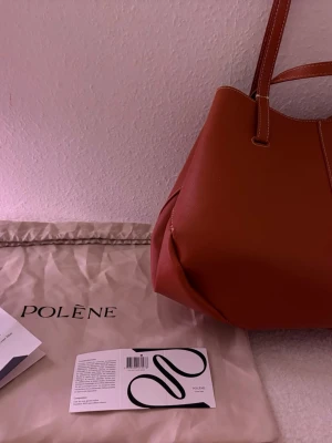 Polène brun handväska i läder - Snygg handväska från Polène i en varm brun färg. Väskan är tillverkad i strukturerat läder och har en modern, mjuk form med dubbla handtag. Medföljer original dustbag och instruktionshäfte. Perfekt för dig som gillar stilrena accessoarer.