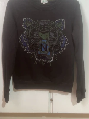 Kenzo tröja - Svart sweatshirt från Kenzo med ikoniskt broderat tigerhuvud i grönt, blått och lila på bröstet. Rund halsringning och långa ärmar. Perfekt statement-plagg för dig som gillar streetwear och vill sticka ut. Fick den som present men var för liten på mig. Priset kan diskuteras.