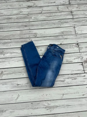 Replay jeans - Säljer ett par blå slim fit jeans från Replay med snygga slitningar och klassisk femficksdesign. Jeansen har dragkedja och knapp framtill samt detaljerade sömmar. Perfekt för dig som gillar en modern och stilren look.