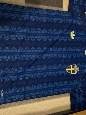 Blå Sverige fotbollströja Adidas - Säljer en blå fotbollströja från Adidas med Sveriges landslagsemblem på bröstet. Tröjan har ett unikt mönster i ljusblått, gula Adidas-detaljer och rund gul krage. Perfekt för dig som vill sticka ut på planen eller heja på Sverige.