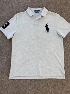 Grå pikétröja från Polo Ralph Lauren - Klassisk grå pikétröja från Polo Ralph Lauren med stor svart broderad logga på bröstet och siffran 3 på ärmen. Tröjan har krage, knappar vid halsen och är tillverkad i mjuk bomull. Perfekt för en avslappnad men stilren look.