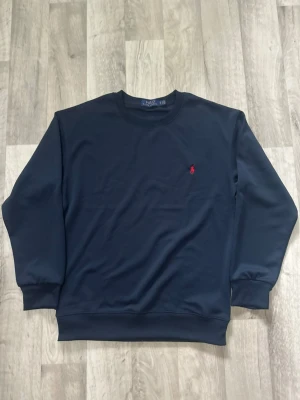 Mörkblå sweatshirt från Ralph Lauren - Klassisk mörkblå sweatshirt från Ralph Lauren med rund halsringning och diskret röd logobrodyr på bröstet. Tröjan har ribbade muddar vid ärmslut och nederkant. Perfekt för en clean och stilren look.