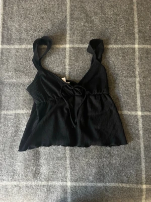 Svart ribbad linnetopp med knyt -  Säljer ett väldigt gulligt men ändå snyggt linne med knytning framtill. Säljer detta linnet då det tyvärr inte har kommit till användning. 