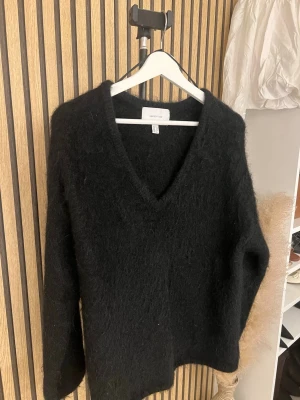 Svart fluffig stickad tröja i mohairmix - Mysig svart stickad tröja från H&M Edition med fluffig yta och djup V-ringning. Tröjan har lång ärm och avslappnad passform, perfekt för lager på lager. Materialet ser ut att vara en mjuk mix med mohair eller liknande.
