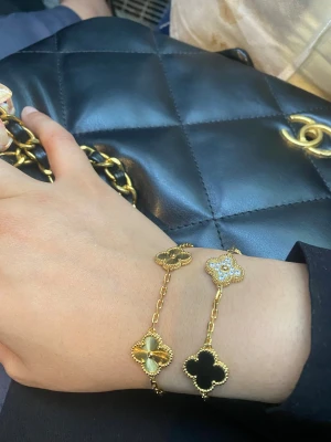Hög kvalitet 18K guldpläterad armband med blommotiv - Snyggt armband i guldton med flera fyrklöverformade hängen, varav ett är dekorerat med glittrande stenar och de andra i svart och guld. Kedjan är tunn och elegant, perfekt för att ge en lyxig touch till din stil. Pris per armband