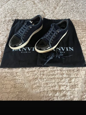 Lanvin mörkblå sneakers med lack - Snygga mörkblå sneakers från Lanvin med vit sula och svart lackad tå. Skorna har klassisk låg modell, mocka och textilmaterial samt matchande mörkblå skosnören. Perfekt för dig som vill ha en exklusiv och stilren look.