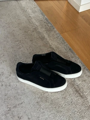 Svarta sneakers från Axel Arigato - Snygga svarta sneakers från Axel Arigato med vit sula och diskret guldfärgad logga på sidan. Skorna har en stilren design med bred resår över vristen istället för klassiska snören. Ovandelen är i mocka vilket ger en lyxig känsla.Mockan är inte alls förstörd dem är som nya eftersom jag bara använt dem till vissa tillfällen 