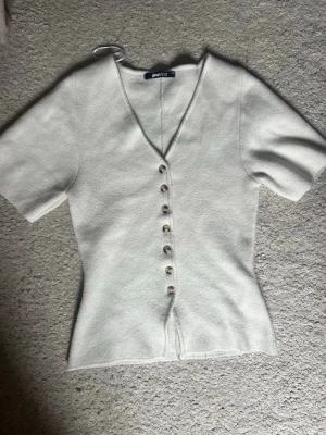 Vit v-ringad topp från Gina Tricot - Snygg vit topp från Gina Tricot med v-ringning och korta ärmar. Toppen har en figurnära passform och knäpps framtill med dekorativa knappar. Perfekt att styla med jeans eller kjol för en clean look.