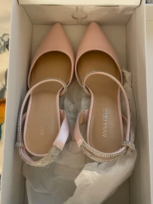 Ljusa rosa pumps med strass från Anna Field - Säljer ett par ljusa rosa pumps från Anna Field med spetsig tå och bred klack. Skorna har en elegant rem med strassdetaljer som ger extra glans. Materialet är satinliknande och klacken är stabil och bekväm. Perfekt för dig som vill ha en chic look. de är helt oanvända fick hem de igår men passade tyvärr inte 