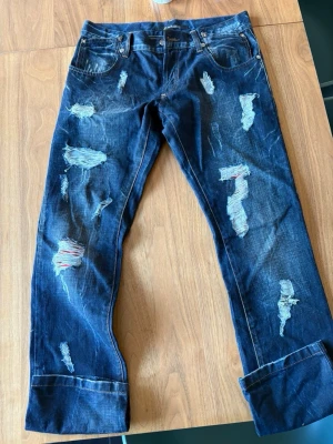 Blå slitna jeans med patchdetaljer - Säljer ett par mörkblå jeans med flera slitningar och patchdetaljer i vitt och rött. Jeansen har raka ben, fem fickor och klassisk knappgylf. Materialet är denim i bomull och de har en avslappnad, trendig look.