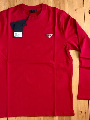 Röd långärmad topp från Prada - Snygg röd långärmad topp från Prada med rund halsringning och ikonisk logga på bröstet. Tillverkad i mjuk bomull som känns skön mot huden. Perfekt för dig som vill ha en enkel men exklusiv look.