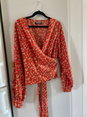 Blommig omlottblus från Fashion Union - Snygg omlottblus från Fashion Union i en röd nyans med små vita blommor och gröna blad. Blusen har långa ärmar med puff och knytband i midjan. Perfekt för dig som gillar en feminin och trendig stil. Materialet känns lätt och luftigt.