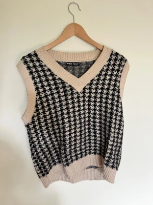 Beige och svart väst från SHEIN - Stickad väst från SHEIN i beige och svart med klassiskt hundtandsmönster. Djup v-ringning och ribbade kanter vid hals, ärmhål och nederkant. Perfekt att bära över en skjorta eller t-shirt för en trendig lager-på-lager-look.