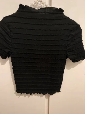 Svart ribbad topp från Hm - Säljer en svart ribbad topp från Divided i storlek XS. Toppen har korta ärmar, croppad passform och en lätt rynkad struktur som ger en cool look. Perfekt att matcha med höga jeans eller kjol.
