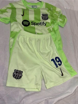 Barcelona matchställ Lamine Yamal #19 - Snyggt Barcelona matchställ i ljusgrönt med tröja och shorts. Tröjan har tryck med Spotify-logga, klubbmärke och Lamine Yamal #19 på ryggen. Shorts med klubbmärke och nummer 19. Tillverkad i lätt och ventilerande polyester, perfekt för fotbollsträning.