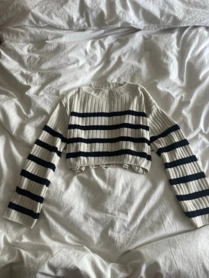 Randig cropped stickad tröja - Säljer en ribbstickad cropped tröja i vitt med svarta horisontella ränder. Tröjan har långa ärmar och rund halsringning. Perfekt för dig som gillar en trendig och avslappnad stil.