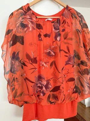 Röd blommig chiffongblus - Snygg röd blus i luftig chiffong med stora blommönster i lila och rosa toner. Blusen har rund halsringning och ett lager av transparent tyg över ett fastsytt underlinne. Perfekt för dig som gillar färg och mönster.