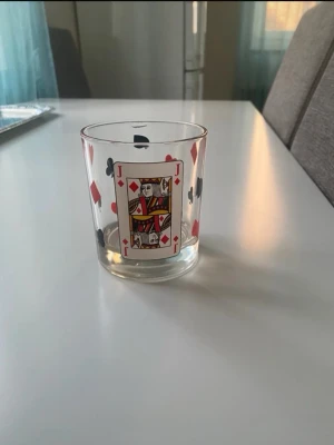 Glas med spelkortsmotiv - Unikt glas med tryck av spelkort, inklusive hjärter, ruter, klöver och spader. På framsidan finns en dam i ruter. Glaset är genomskinligt och dekorerat med färgglada symboler, perfekt för dig som gillar kortspel eller vill ha något annorlunda i köksskåpet.