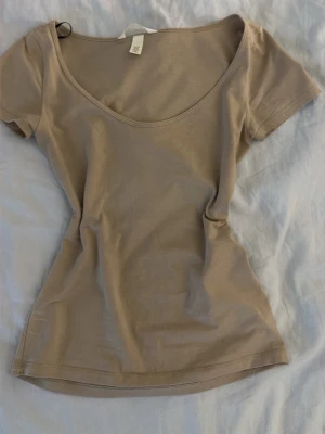 Beige t-shirt med djup ringning - En enkel och stilren beige t-shirt från H&M med djup rundringning och korta ärmar. Tillverkad i mjuk bomull som känns skön mot huden. Perfekt basplagg att matcha med allt i garderoben.
