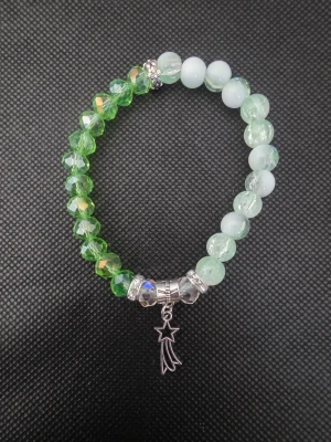 Handgjort gröntoch vitt pärlarmband med stjärnhänge - Snyggt handgjort armband med glittrande gröna och vita pärlor i olika nyanser. Armbandet har ett silverfärgat hänge i form av en stjärna med svans och dekorativa metallpärlor. Perfekt accessoar för att lyfta din stil.
