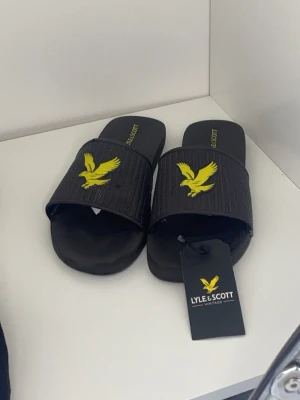 Tofflor -   Märke: Lyle & Scott (Vintage)  Typ: Tofflor / Slides / Badtofflor  Färg: Svarta med märkets klassiska gula örn-logotyp på ovansidan samt gult tryck på sulan  Detaljer: Mönstrad textur på ovansidan, helt nya med prislapp/tagg kvar  Tillbehör: En transparent originalplastpåse med Lyle & Scott-tryck och varningsinformation (används för förvaring/upphängning) pris kan diskuteras 