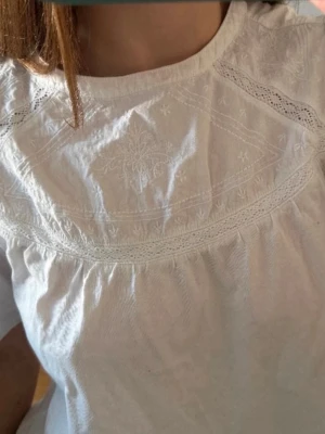 Vit blus med broderi från Zara - Söt vit blus från Zara med fina broderade detaljer och spets längs axlar och bröst. Blusen har rund halsringning och korta ärmar. Perfekt för en fräsch och stilren look. Tillverkad i mjukt bomullstyg.