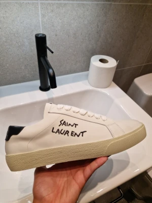 Saint Laurent skor - [Helt nya i storlek 39. Pris: 1400💸 men kan diskuteras vid snabb affär🚀]                     Snygga vita sneakers från Saint Laurent med handskriven logga på sidan. Skorna har en klassisk låg siluett, vita skosnören och svart detalj på hälen. Tillverkade i slätt läder med en beige sula som ger en clean och trendig look.         