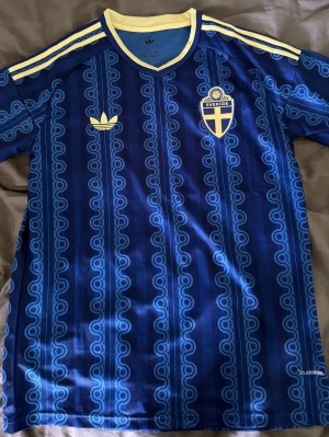 Blå Sverige fotbollströja Adidas - Säljer en blå fotbollströja från Adidas med Sveriges landslagsemblem och gula detaljer. Tröjan har ett retroinspirerat mönster, gula ränder på axlarna och gul krage. Tillverkad i lätt och ventilerande Climalite-material, perfekt för träning eller match.