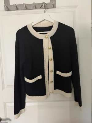 Svart kofta med vita detaljer - Stilren svart kofta med kontrasterande vita kanter och guldfärgade knappar. Rund halsringning och två fickor framtill. Perfekt för en chic och klassisk look. Mjuk och skön stickad kvalitet.
